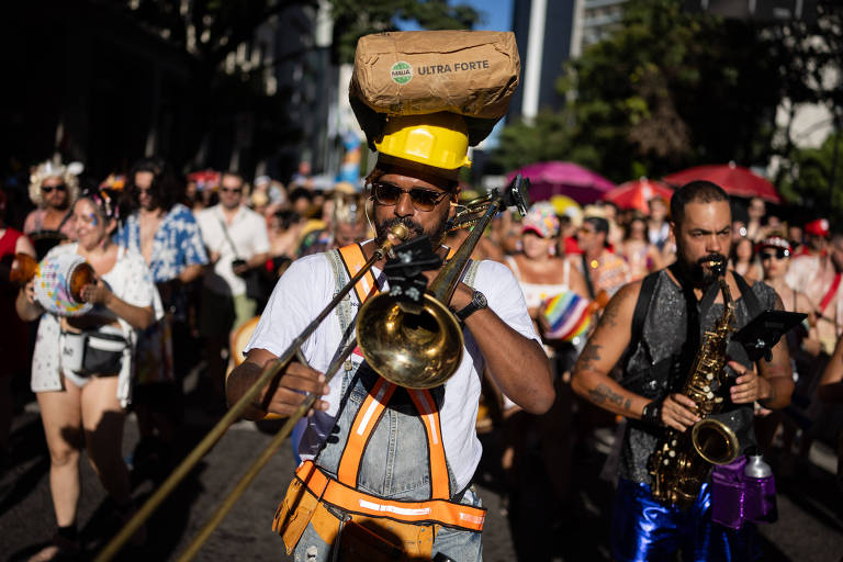 A imagem mostra um desfile de Carnaval com uma multidão ao fundo. Em primeiro plano， um músico fantasiado toca trombone， usando um macacão laranja e uma camiseta branca. Ao lado dele， outro músico toca saxofone. A multidão é colorida， com pessoas usando roupas festivas.