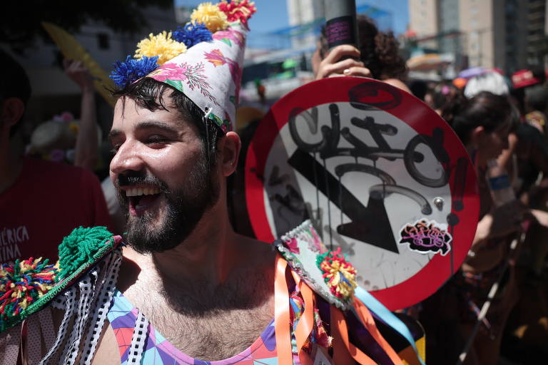 Um homem sorridente participa de uma festa de Carnaval, usando um traje colorido com pompons e um chapéu de festa. Ele está segurando um sinal de trânsito que indica uma direção, que está grafitado. Ao fundo, há outras pessoas e um ambiente festivo.