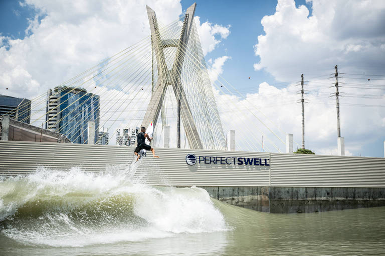 A imagem mostra um surfista realizando uma manobra em uma onda em um ambiente urbano. Ao fundo， há uma estrutura arquitetônica moderna com cabos e pilares， além de edifícios altos. O céu está parcialmente nublado， com nuvens brancas e azuis. Na parte inferior da imagem， há uma placa com a inscrição 039;PERFECT SWELL039;.