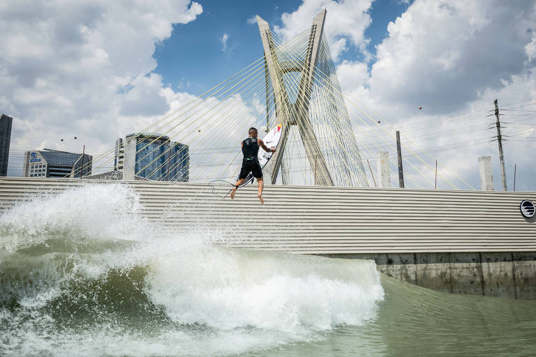 Um surfista está realizando uma manobra sobre uma onda em um ambiente urbano. Ao fundo， é possível ver um grande edifício e uma ponte com estruturas modernas. O céu está parcialmente nublado， com algumas nuvens brancas e azuis. A água está agitada， criando uma onda significativa.