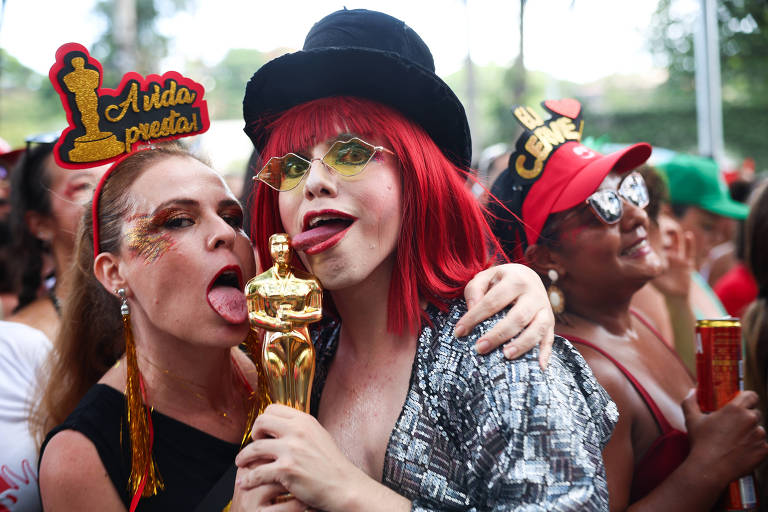 A imagem mostra um grupo de pessoas em um evento festivo, possivelmente durante o Carnaval. Duas mulheres estão em destaque, uma com cabelo loiro e uma tiara que diz 'Aí, dá preta', e a outra com cabelo vermelho, óculos coloridos e segurando uma estatueta dourada. Ao fundo, outras pessoas também estão se divertindo, algumas usando chapéus e acessórios coloridos.