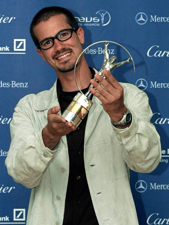 Um homem， que é Bob Burnquist， segura um troféu em uma cerimônia de premiação. Ele usa óculos e uma camisa preta sob um casaco claro. O fundo é azul com logotipos da Mercedes-Benz e do Laureus Sports Awards