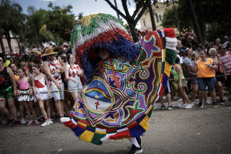 A imagem mostra uma pessoa vestida com uma fantasia colorida e elaborada, que possui um grande chapéu decorado com franjas coloridas. A fantasia é adornada com desenhos vibrantes, incluindo um olho e uma cruz. Ao fundo, há uma multidão de pessoas assistindo ao desfile, algumas vestindo roupas de festa e outras com fantasias. O ambiente é festivo, com árvores e edifícios visíveis.