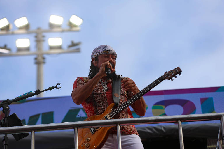 Um artista está se apresentando em um palco, segurando um microfone com a mão esquerda e uma guitarra com a mão direita. Ele usa uma camisa colorida e um lenço na cabeça. Ao fundo, há luzes de palco acesas e uma faixa com cores vibrantes.