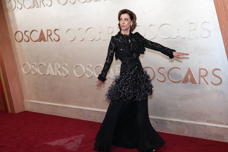 Uma mulher está posando em um tapete vermelho em um evento do Oscar. Ela usa um vestido preto com detalhes em brilho e uma saia volumosa. Ao fundo， há uma parede com a palavra 039;OSCARS039; repetida em letras grandes.