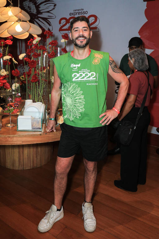 Um homem está posando em um evento. Ele usa uma camiseta verde sem mangas com a inscrição '2222' e um desenho floral. Ele está com shorts pretos e tênis brancos. Ao fundo, há uma decoração com flores vermelhas e outras pessoas visíveis.