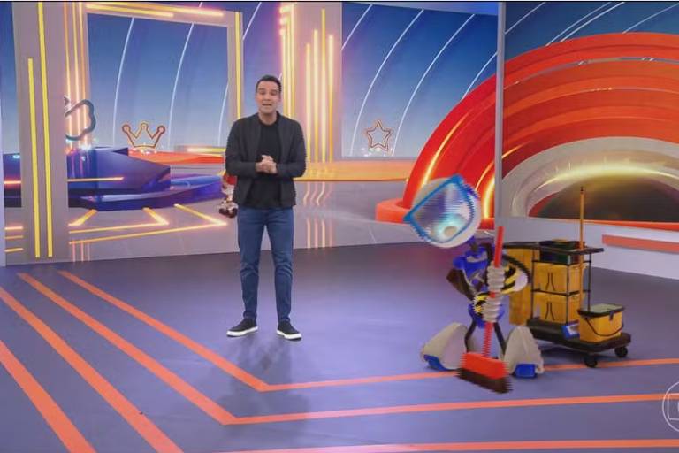 Um apresentador de pé em um estúdio de televisão, ao lado de um robô que está limpando o chão com um equipamento de limpeza. O estúdio tem um fundo colorido com arcos vermelhos e elementos gráficos. O apresentador está vestindo uma camiseta preta e calças jeans, segurando um controle remoto.