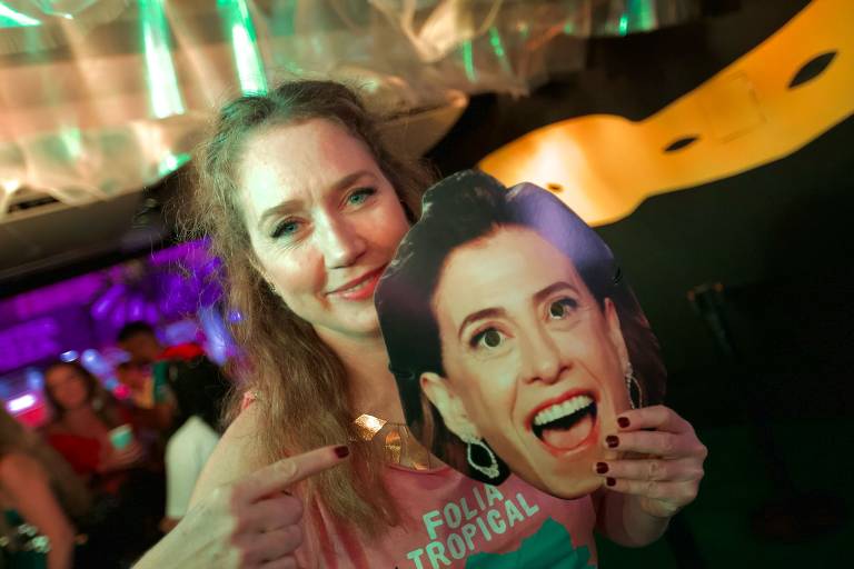 Uma mulher está em uma festa, segurando uma máscara de papel com o rosto de outra pessoa, que está sorrindo. Ela usa uma camiseta rosa com a frase 'FOLIA TROPICAL' e está apontando para a máscara. O ambiente é festivo, com luzes coloridas ao fundo e outras pessoas visíveis.