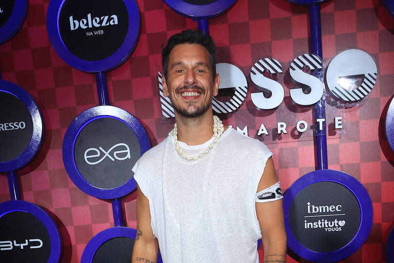 Um homem está posando em um evento, vestindo uma camiseta branca e calças brancas. Ele sorri e está em frente a um fundo vermelho com círculos pretos que contêm logotipos de marcas. O ambiente parece ser festivo e bem iluminado.
