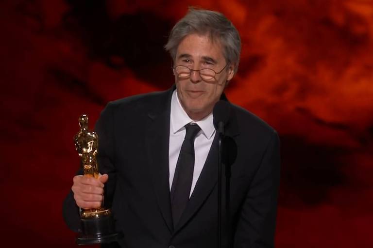 Um homem de cabelos grisalhos， usando óculos e um terno escuro， está em pé segurando um Oscar em uma mão. Ele está em um palco com um fundo de cor laranja e uma placa na frente que diz 039;I039;M STILL HERE039;， 039;BRAZIL039;， e 039;INTERNATIONAL FEATURE FILM039;.
