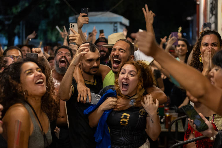 A imagem mostra um grupo de pessoas em um ambiente festivo, todas expressando alegria e entusiasmo. Algumas pessoas estão sorrindo e gritando, enquanto outras se abraçam. O cenário parece ser uma festa ou uma comemoração, com bebidas visíveis nas mesas ao fundo. A iluminação é suave, sugerindo que é à noite.