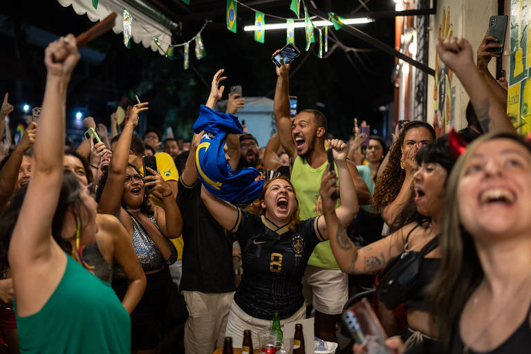 Uma multidão animada em um bar， com pessoas levantando os braços e gritando de alegria. Algumas pessoas estão vestindo camisetas de futebol， enquanto outras seguram celulares. O ambiente é festivo， com bandeirinhas decorativas e uma atmosfera de celebração.