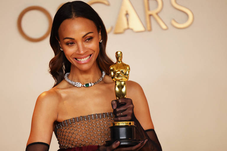 Uma mulher sorridente segura uma estatueta do Oscar em um evento. Ela está vestida com um vestido elegante， com detalhes em brilho e uma joia ao redor do pescoço. O fundo apresenta a palavra 039;OSCARS039; em letras grandes e douradas.