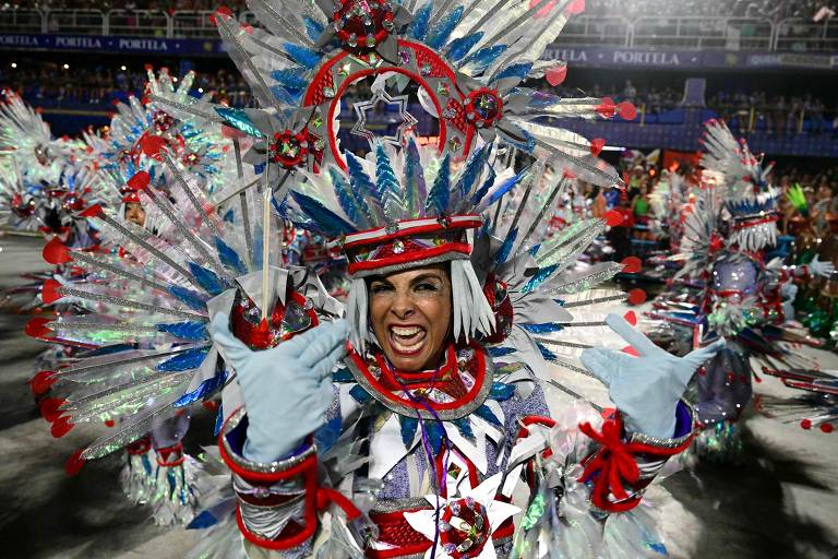 A imagem mostra uma mulher em um traje de carnaval vibrante, com penas e brilhos, dançando e fazendo um gesto de rock com as mãos. Ela tem um sorriso expressivo e está cercada por outras pessoas em trajes semelhantes, todos em um ambiente festivo.