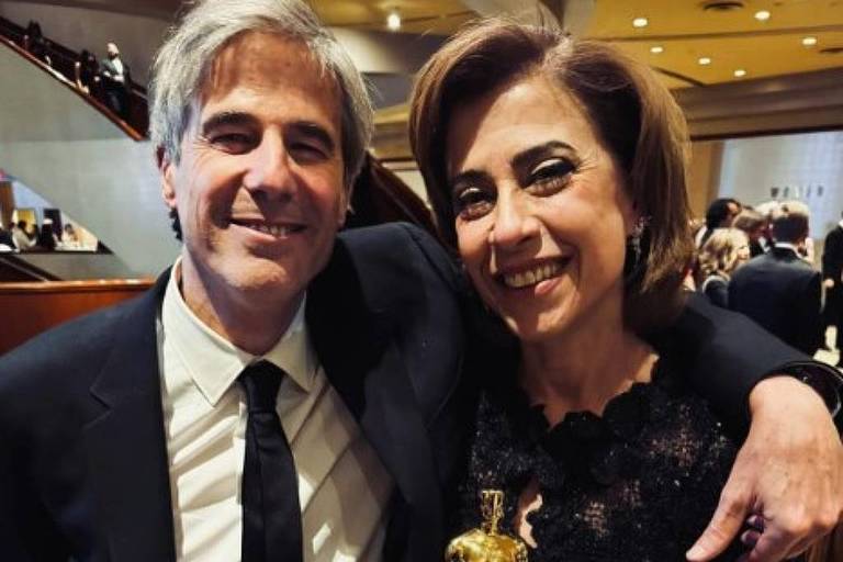 Um homem e uma mulher posam juntos em um evento， ambos sorrindo. O homem está vestindo um terno preto com uma gravata preta e a mulher está usando um vestido preto com detalhes em penas. Ela segura uma estatueta dourada， possivelmente um prêmio， enquanto o homem a abraça levemente.