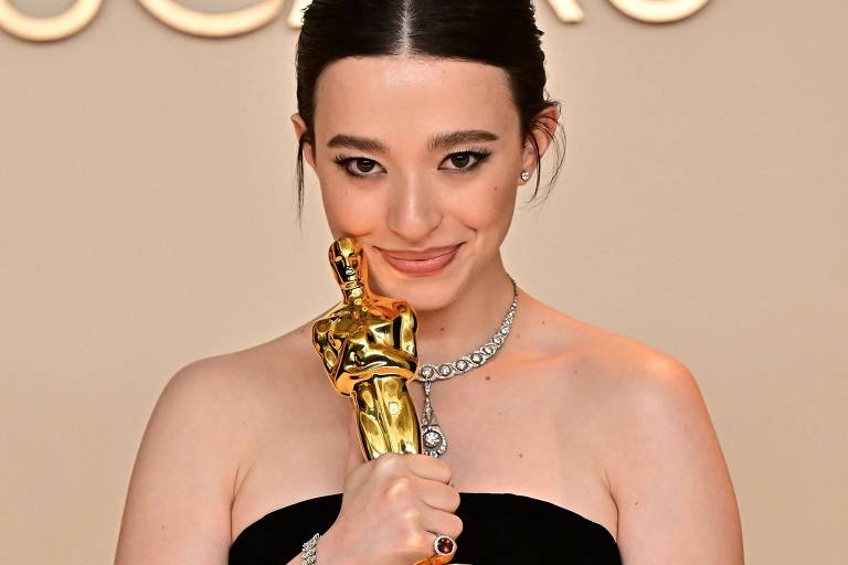 Uma mulher segurando uma estatueta do Oscar， com um vestido preto e joias brilhantes. Ela está sorrindo levemente e olhando para a câmera， com o fundo em tom neutro.
