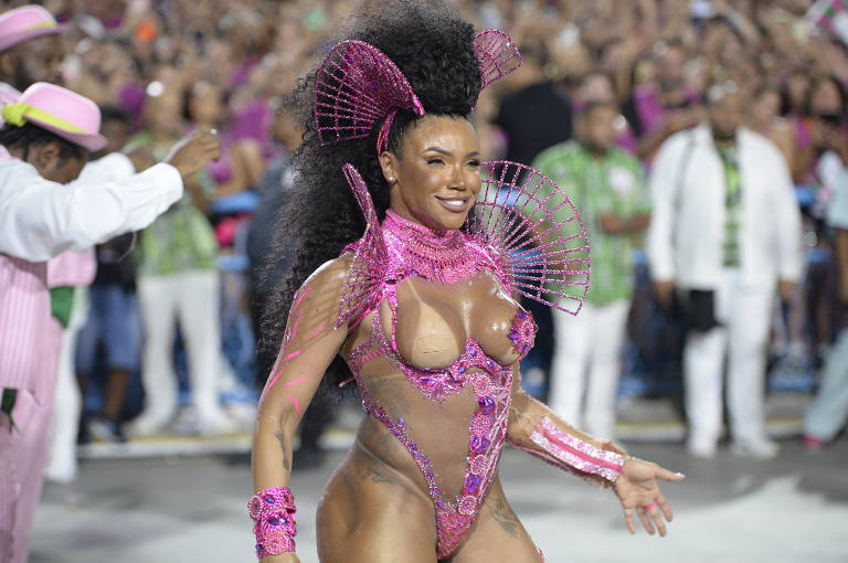 Uma mulher com cabelo volumoso e uma fantasia brilhante de cor rosa, que destaca seu corpo, está desfilando em um evento de carnaval. Ao fundo, há uma multidão de pessoas vestidas em roupas coloridas, algumas com chapéus e acessórios. A atmosfera é festiva e vibrante.
