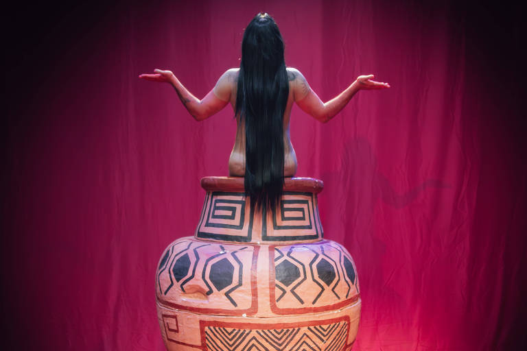 A imagem mostra uma figura feminina de costas, com longos cabelos pretos, sentada em um grande vaso decorativo. O vaso possui padrões geométricos em tons de laranja e preto. A figura está com os braços levantados, em um gesto que sugere abertura ou acolhimento. O fundo é de um tom vermelho, criando um contraste com o vaso e a figura.