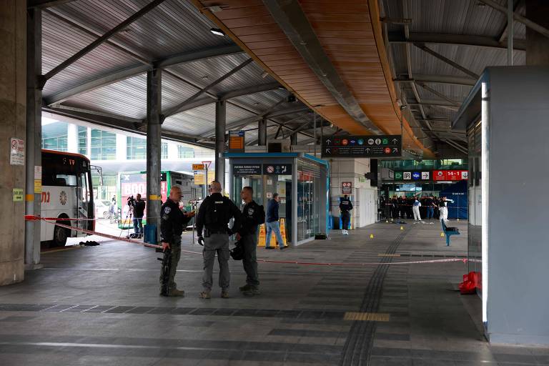 A imagem mostra o interior de uma estação de ônibus. Há dois policiais em primeiro plano, conversando entre si. Ao fundo, é possível ver um ônibus estacionado e algumas pessoas. A área está cercada com fita de isolamento, indicando uma possível investigação. O teto é alto e a iluminação é artificial, com painéis informativos visíveis na parte superior.