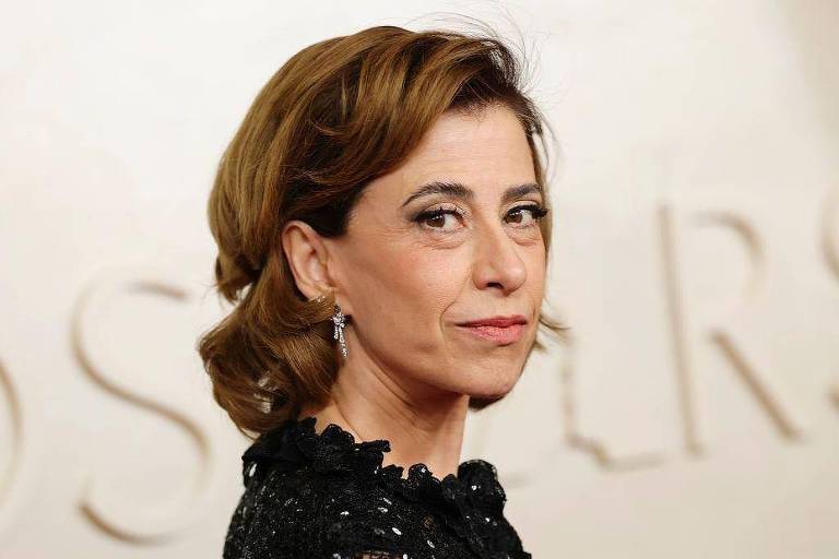 A imagem mostra uma mulher com cabelo castanho claro, preso em um coque, usando um vestido preto com detalhes. Ela está olhando para o lado, com uma expressão séria. O fundo é desfocado, mas parece ter a palavra 'OSCARS' em destaque.