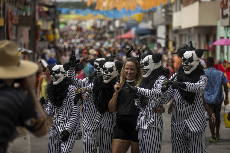 A imagem mostra uma rua movimentada durante um desfile de Carnaval. No primeiro plano, uma mulher sorridente está posando para a câmera, enquanto atrás dela há um grupo de palhaços com máscaras brancas e roupas listradas em preto e branco. O fundo é composto por uma multidão de pessoas e bandeirinhas coloridas penduradas, criando um ambiente festivo.