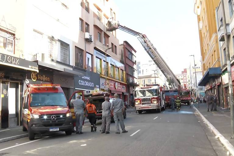 A imagem mostra uma rua com várias viaturas de bombeiros estacionadas. Há uma escada de bombeiros estendida em direção a um andar superior de um edifício. Algumas pessoas， incluindo bombeiros， estão na calçada observando a situação. O ambiente urbano é composto por lojas e edifícios ao fundo.