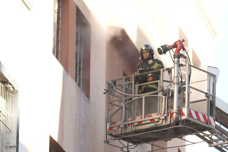 Um bombeiro está em uma plataforma elevatória， combatendo um incêndio em um prédio. O bombeiro está vestido com equipamento de proteção e utiliza uma mangueira para direcionar água para o local em chamas. Há fumaça saindo de uma janela do edifício.