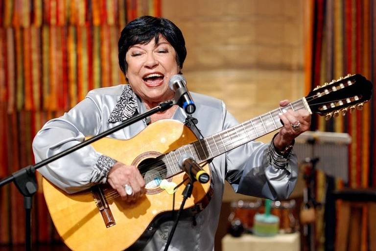 Uma mulher de cabelos curtos e escuros， vestindo uma blusa prateada， está cantando e tocando violão em um palco. Ela sorri amplamente enquanto segura o microfone com uma mão e o violão com a outra. Ao fundo， há uma decoração colorida com faixas verticais e instrumentos musicais visíveis.