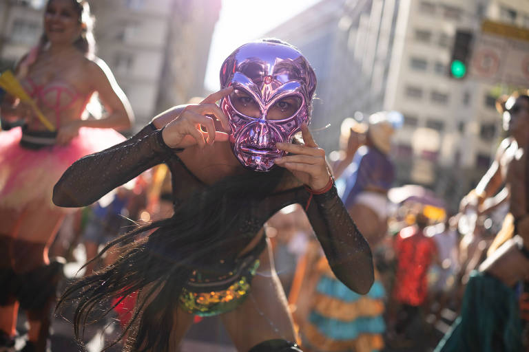 A imagem mostra uma pessoa em um desfile de Carnaval no Rio de Janeiro， usando uma máscara roxa brilhante e roupas coloridas. A pessoa está em primeiro plano， fazendo um gesto com as mãos， enquanto ao fundo há outras pessoas vestidas com trajes festivos e coloridos， em um ambiente urbano iluminado pelo sol.