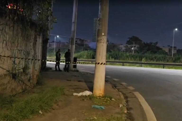 A imagem mostra uma cena noturna em uma área urbana. Há um muro à esquerda coberto de grafites e uma calçada com grama. Dois homens estão em pé próximos a um poste de luz， que está iluminado. A estrada à direita é pavimentada e há uma faixa de pedestres visível. Ao fundo， é possível ver algumas edificações e vegetação. A iluminação é fraca， criando uma atmosfera sombria.
