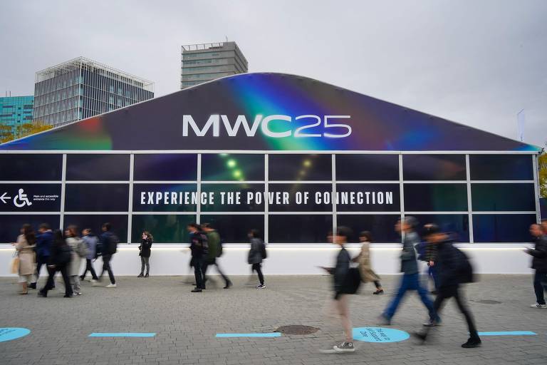 A imagem mostra a entrada do evento MWC 25, com uma estrutura grande e moderna. Na fachada, está escrito 'MWC25' e 'EXPERIENCE THE POWER OF CONNECTION'. Há várias pessoas caminhando em frente à entrada, e o céu está nublado.
