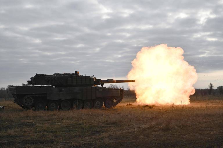 A imagem mostra um tanque de guerra disparando， com uma explosão visível na extremidade do canhão. O cenário é um campo aberto com grama seca e um céu nublado ao fundo.