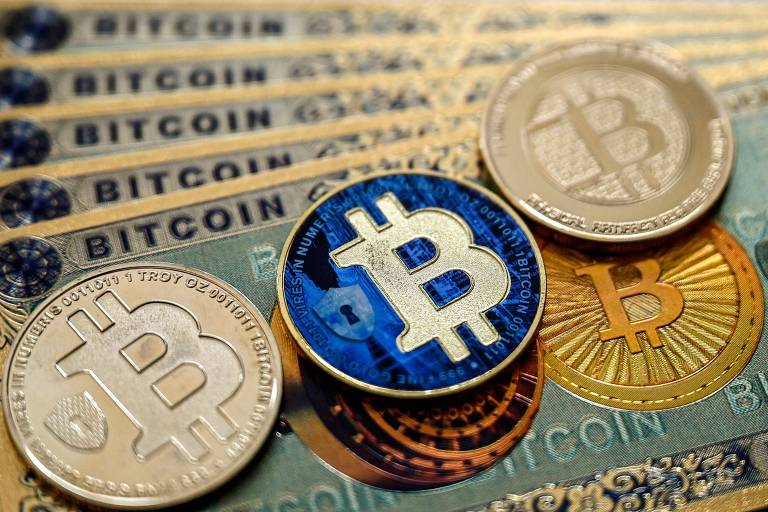A imagem mostra várias notas de Bitcoin empilhadas， com três moedas de Bitcoin em cima. As notas têm a palavra 039;BITCOIN039; impressa e as moedas apresentam o símbolo do Bitcoin em diferentes designs， incluindo uma moeda dourada， uma azul e uma prateada.