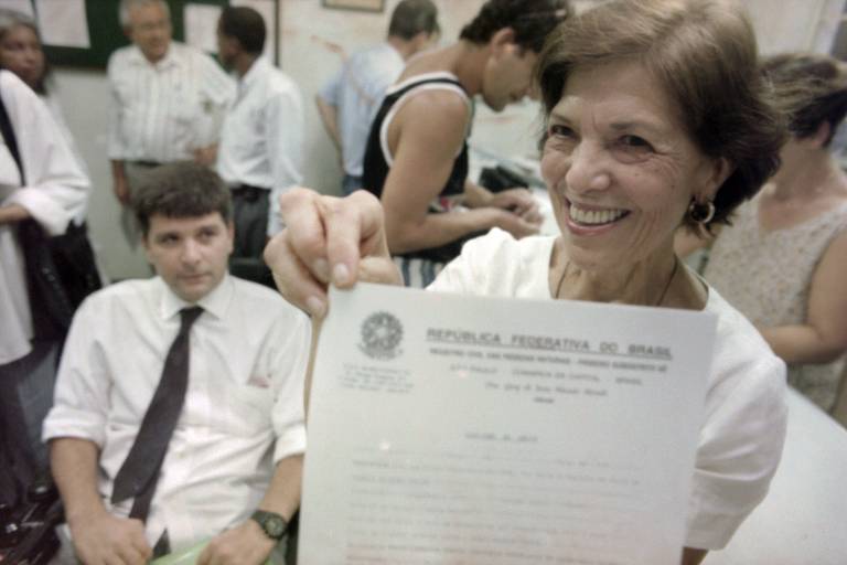 A imagem mostra uma mulher sorridente segurando um documento em primeiro plano. Ela está em um ambiente interno， possivelmente um escritório ou uma repartição pública. Ao fundo， há outras pessoas， incluindo um homem sentado à esquerda， que parece estar concentrado em algo. O documento que a mulher segura tem o selo da República Federativa do Brasil.