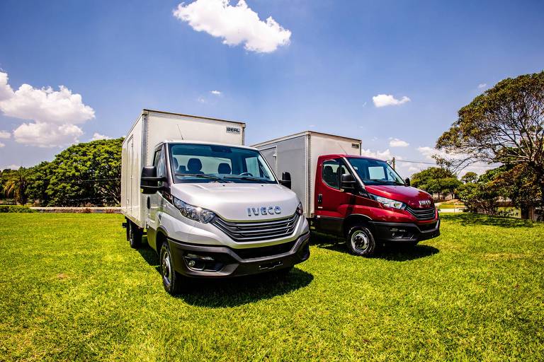 A imagem mostra dois caminhões estacionados em um campo verde. O caminhão à esquerda é de cor prata e possui uma carroceria branca. O caminhão à direita é de cor vermelha， também com carroceria branca. O céu está claro com algumas nuvens brancas e há árvores ao fundo.