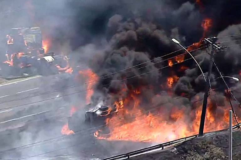 A imagem mostra um incêndio intenso em um caminhão na rodovia Presidente Dutra， no Rio. Chamas altas e fumaça densa se elevam， cobrindo a área ao redor. Linhas de energia estão visíveis， e a cena transmite uma sensação de emergência.