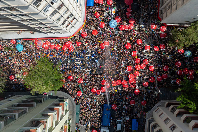 Imagem aérea de uma grande manifestação em uma rua urbana， com uma multidão densa de pessoas. A maioria das pessoas está usando roupas coloridas e há muitos guarda-sóis vermelhos espalhados pela área. Edifícios altos cercam a cena， e algumas árvores podem ser vistas nas bordas.