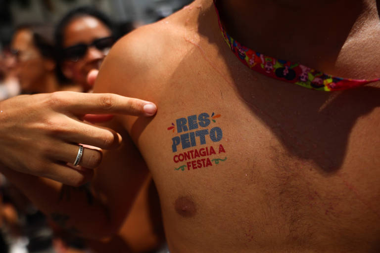 A imagem mostra um homem com um tatuagem no peito que diz 'RESPEITO CONTAGIA A FESTA'. Ele está apontando para a tatuagem com um dedo. Ao fundo, há pessoas em um ambiente festivo, mas não são claramente visíveis.