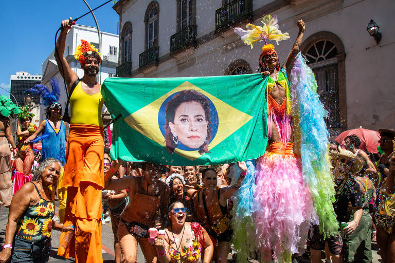 A imagem mostra um grupo de pessoas celebrando durante um carnaval de rua no Brasil. Duas pessoas em trajes coloridos seguram uma grande bandeira do Brasil com uma imagem de uma mulher no centro. Ao fundo, há uma multidão de foliões, alguns usando roupas de festa e outros com acessórios coloridos. O ambiente é festivo, com confetes e uma atmosfera de alegria.