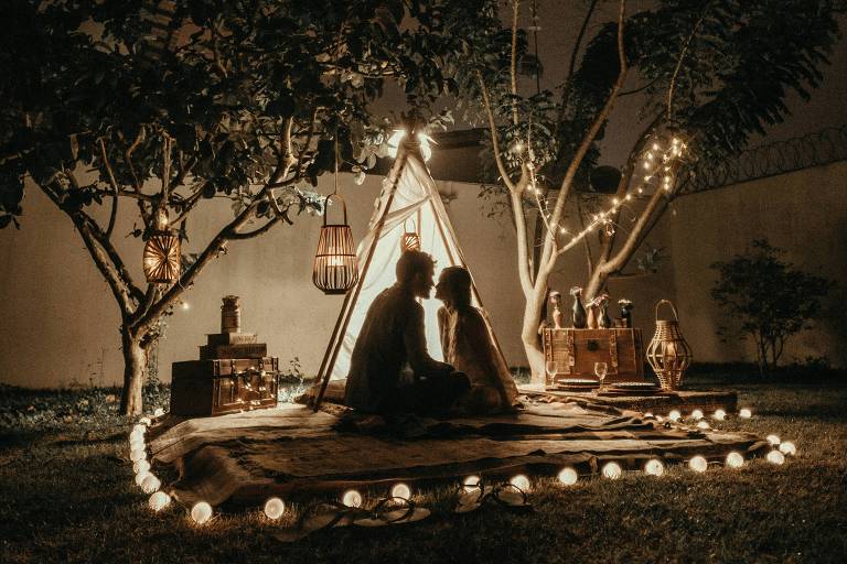 A imagem mostra um casal sentado em um tapete em um jardim à noite, sob uma estrutura de tenda feita de madeira e coberta com luzes. Ao redor, há árvores e uma iluminação suave que destaca o ambiente acolhedor. A cena é em tons escuros, com luzes quentes criando uma atmosfera romântica.