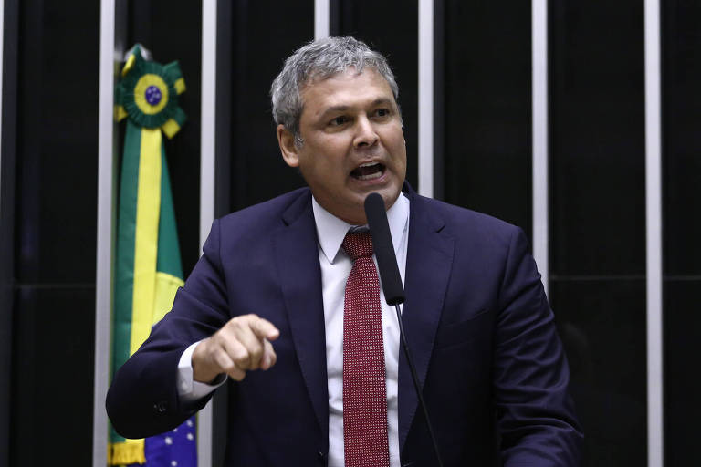 Um homem em um terno escuro e gravata vermelha está falando em um microfone， gesticulando com a mão. Ao fundo， há uma bandeira do Brasil e as paredes do plenário do Parlamento.