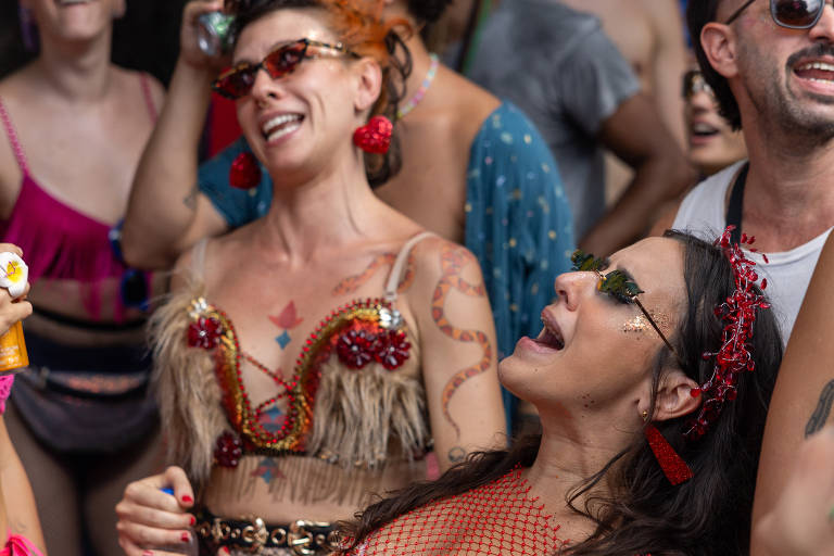 A imagem mostra um grupo de pessoas se divertindo em uma festa. Duas mulheres estão em destaque, uma à esquerda com um top de penas e acessórios coloridos, e outra à direita com um vestido de rede e enfeites no cabelo. Ambas estão sorrindo e dançando, cercadas por outras pessoas que também parecem estar se divertindo. O ambiente é vibrante e festivo.