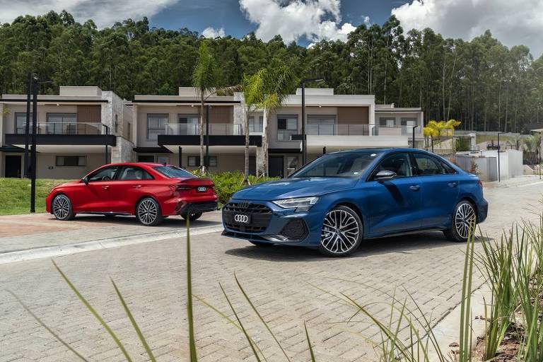 A imagem mostra dois carros estacionados em uma rua de um bairro moderno. Um carro é vermelho e o outro é azul， ambos com design esportivo. Ao fundo， há casas contemporâneas com fachadas em tons neutros e um céu parcialmente nublado. A vegetação ao redor é verde e bem cuidada.