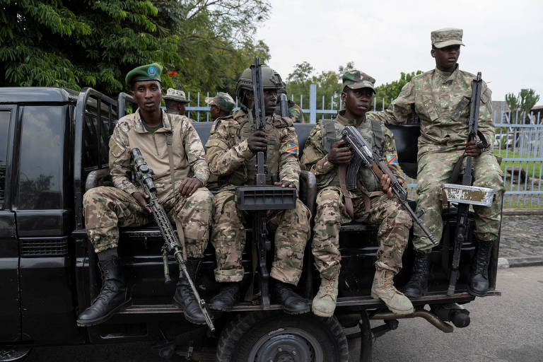 Cinco soldados armados estão sentados na parte de trás de um veículo militar. Eles usam uniformes camuflados e estão em uma posição de vigilância. O ambiente ao fundo é urbano， com uma cerca e vegetação visível.
