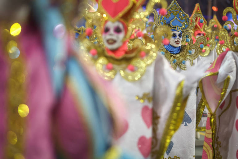 A imagem mostra um grupo de pessoas vestidas com fantasias elaboradas， caracterizadas por máscaras e trajes brancos decorados com detalhes em dourado， rosa e azul. As máscaras têm rostos pintados com expressões alegres e são adornadas com elementos brilhantes e coloridos. O fundo é desfocado， destacando as figuras em primeiro plano， que parecem estar participando de um desfile de Carnaval.