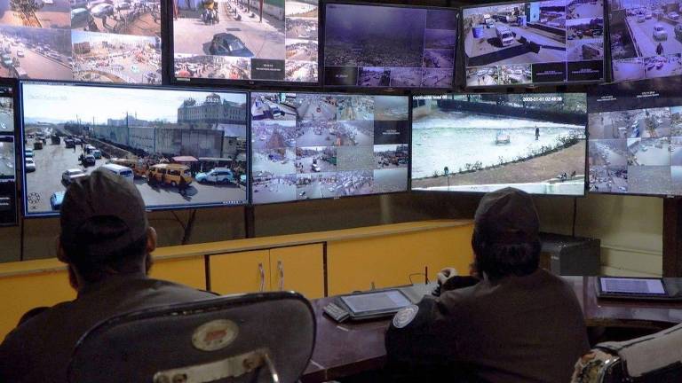A imagem mostra um centro de monitoramento de segurança com várias telas exibindo imagens de câmeras de vigilância. Duas pessoas， aparentemente agentes de segurança， estão sentadas em cadeiras， observando as telas. As imagens nas telas mostram diferentes áreas， incluindo ruas e um corpo d039;água.