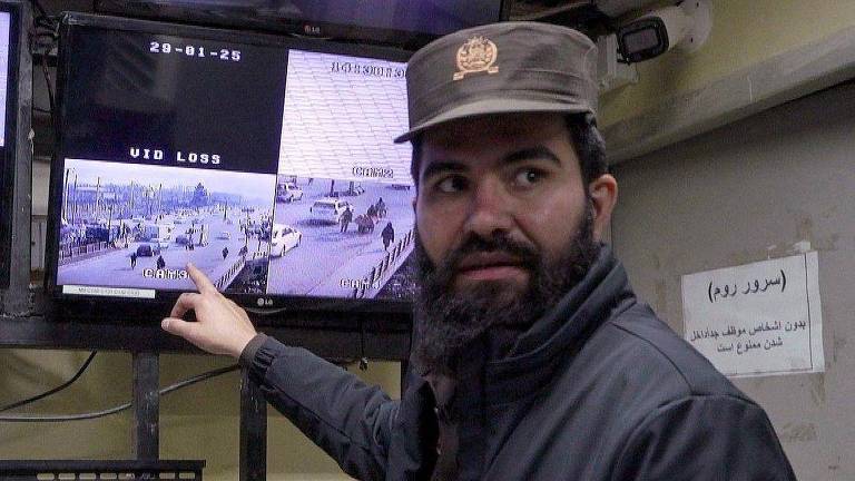 Um homem com barba e usando um boné e uma jaqueta escura está apontando para um monitor de segurança. O monitor exibe várias câmeras de vigilância， algumas mostrando imagens de uma área externa com veículos e pessoas. Há um aviso em uma parede ao fundo， escrito em árabe.