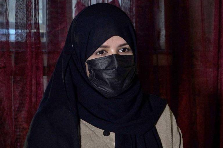 A imagem mostra uma mulher usando um hijab preto e uma máscara facial preta. Ela está em um ambiente interno， com cortinas vermelhas ao fundo. A mulher parece estar em um momento de reflexão ou conversa.