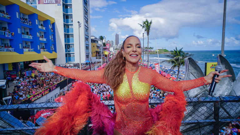 Uma artista se apresenta em um palco ao ar livre, vestindo um traje brilhante com penas coloridas nas mangas e na parte inferior. Ela está sorrindo e com os braços abertos, em frente a uma grande multidão que assiste. Ao fundo, há prédios e o mar, sob um céu parcialmente nublado.