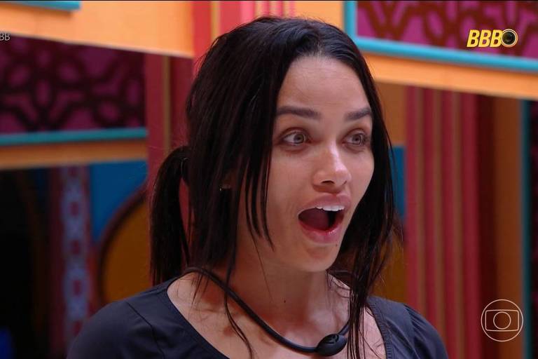 A imagem mostra uma mulher com cabelo longo e escuro, preso em um coque bagunçado, com uma expressão de surpresa. Ela está usando uma blusa preta e parece estar em um ambiente colorido, com paredes decoradas em tons vibrantes. Ao fundo, há elementos decorativos que sugerem um cenário de reality show.